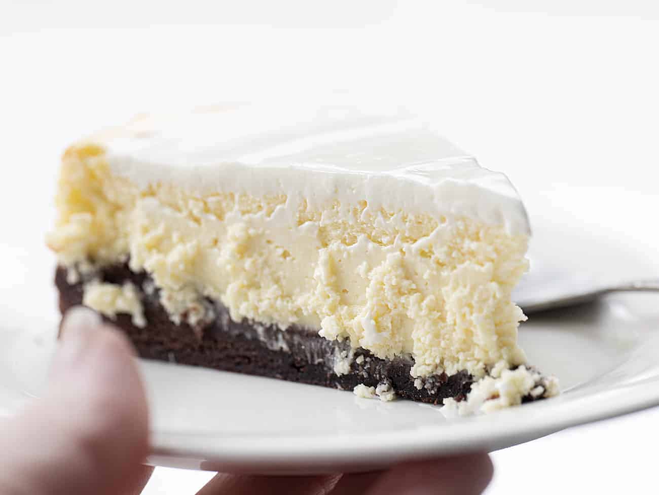 Hand Holding White Plate with Brownie Bottom Cheesecake Recipe with Sour Cream Frosting Topping and Fork Resting Next to It