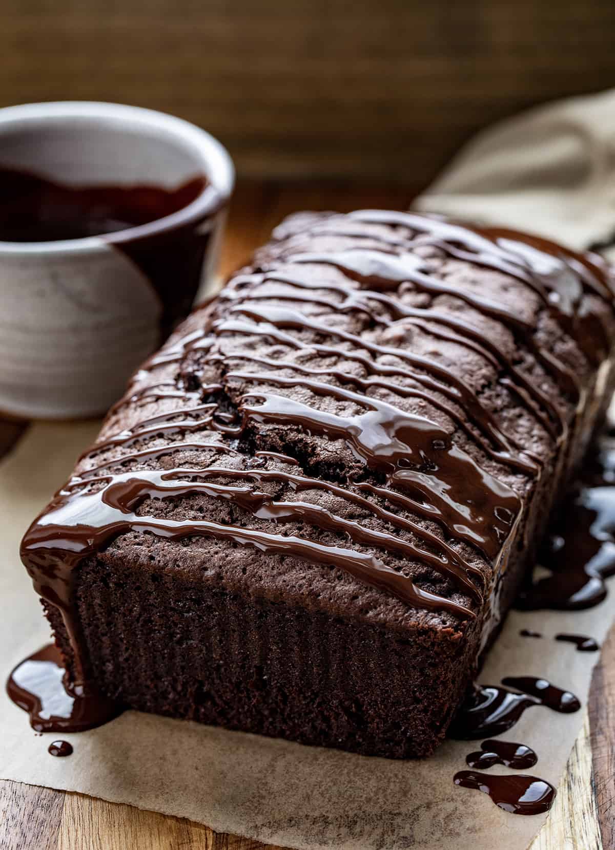 Brownie Bread is a loaf of fudgy chocolate bread that is like a brownie but cut and served like a slice of bread. Then, milk chocolate ganache gives the loaf even more chocolate flavor! It is a quick bread that will quickly be gone, for sure! This delicious treat or dessert will satisfy your sweet tooth and chocolate craving. Brownie Bread on a Cutting Board with Chocolate Drizzled Over Top and a Knife.