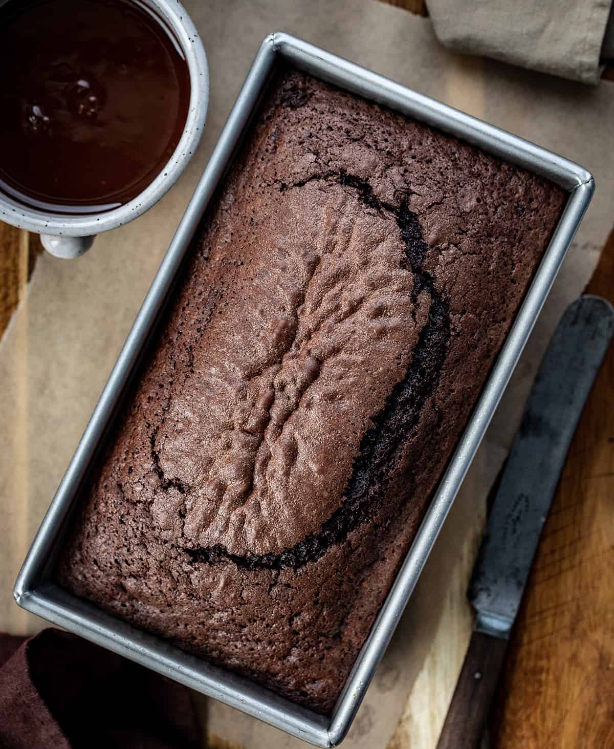 Brownie Bread is a loaf of fudgy chocolate bread that is like a brownie but cut and served like a slice of bread. Then, milk chocolate ganache gives the loaf even more chocolate flavor! It is a quick bread that will quickly be gone, for sure! This delicious treat or dessert will satisfy your sweet tooth and chocolate craving. Overhead Image of Brownie Bread with a Knife and Chocolate Glaze for on Top.
