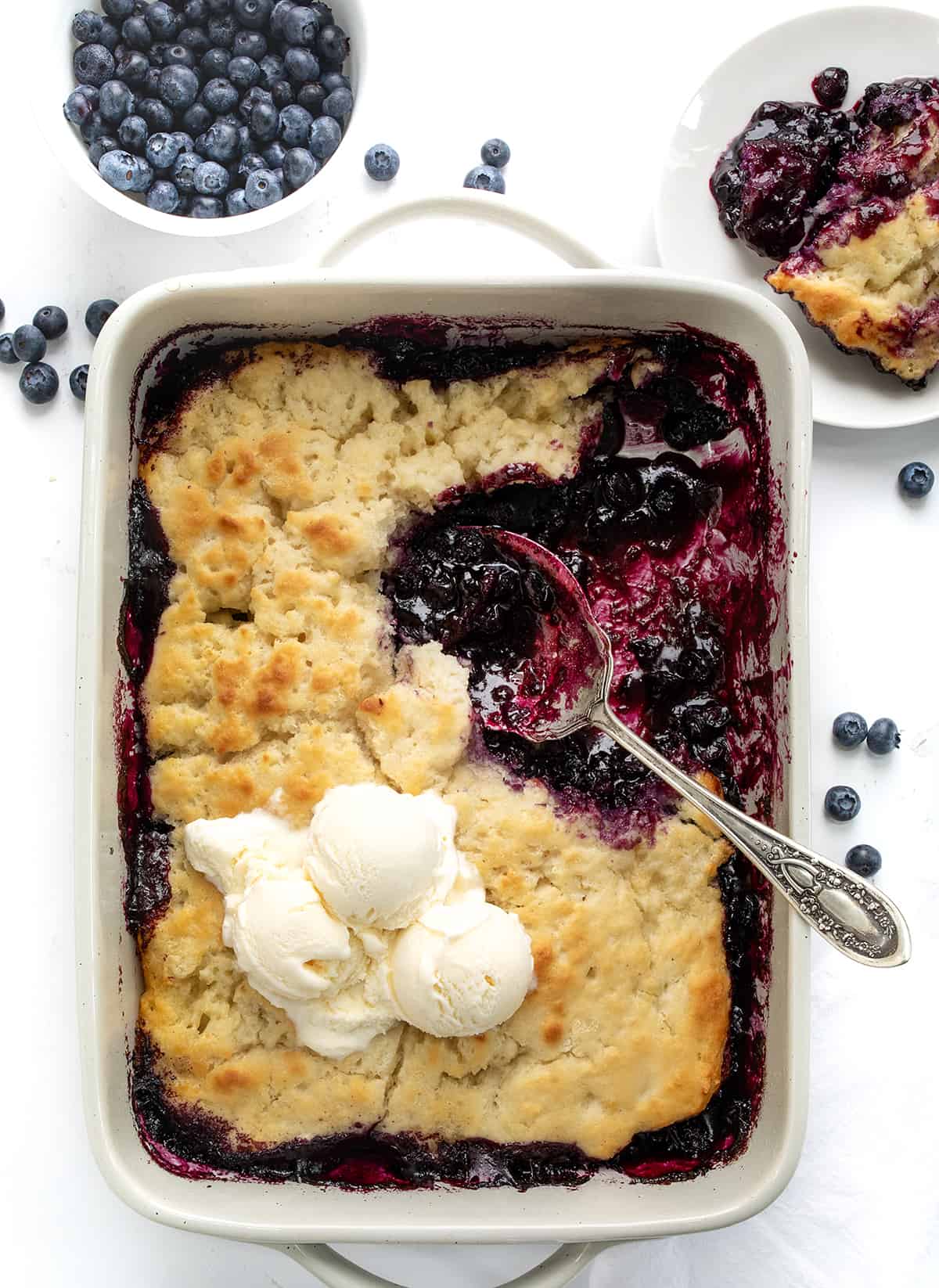 Butter Swim Biscuit Blueberry Cobbler is a delightful dessert that combines homemade blueberry filling with tender, buttery biscuits. It's a perfect treat for gatherings, family meals, or anytime you crave a comforting and fruity dessert. It is so easy to throw together and ready in 30 minutes! Pan of Butter Swim Biscuit Blueberry Cobbler with Ice Cream and a Plated Piece Next to Fresh Blueberries from Overhead.