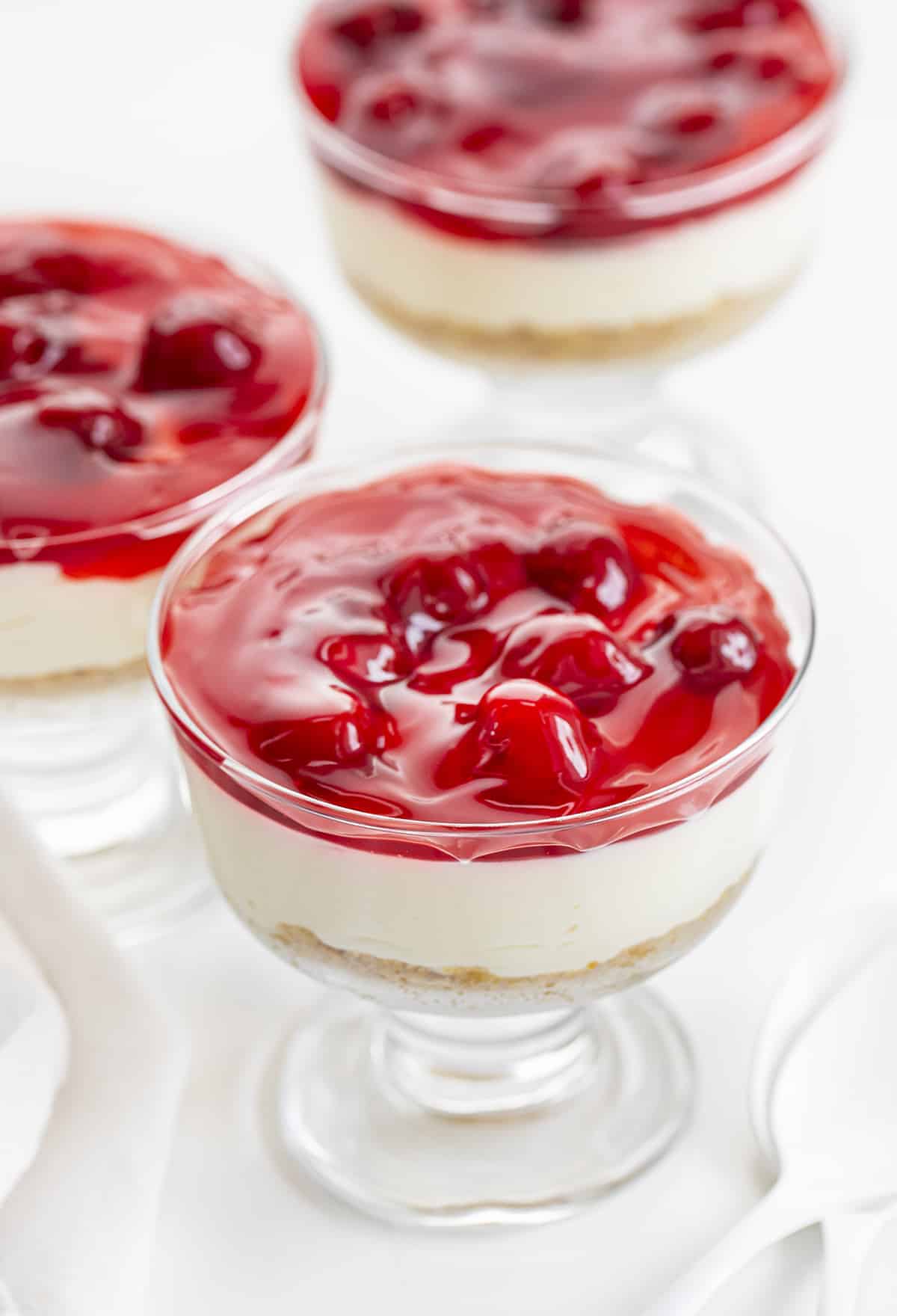 No-Bake Cherry Cheesecake Parfaits are a cheesecake dessert with a graham cracker crust and cherry pie topping, all served in a cup! Dessert, No Bake Dessert, Cherry Cheesecake, Cheesecake Parfait, Cherry Cheesecake Parfait, Cheesecake in a Cup, Creamy Cheesecake, BBQ Dessert, Cookout Dessert, i am baker, iambaker 3 No-Bake Cherry Cheesecake Parfaits on a White Counter with White Spoons.