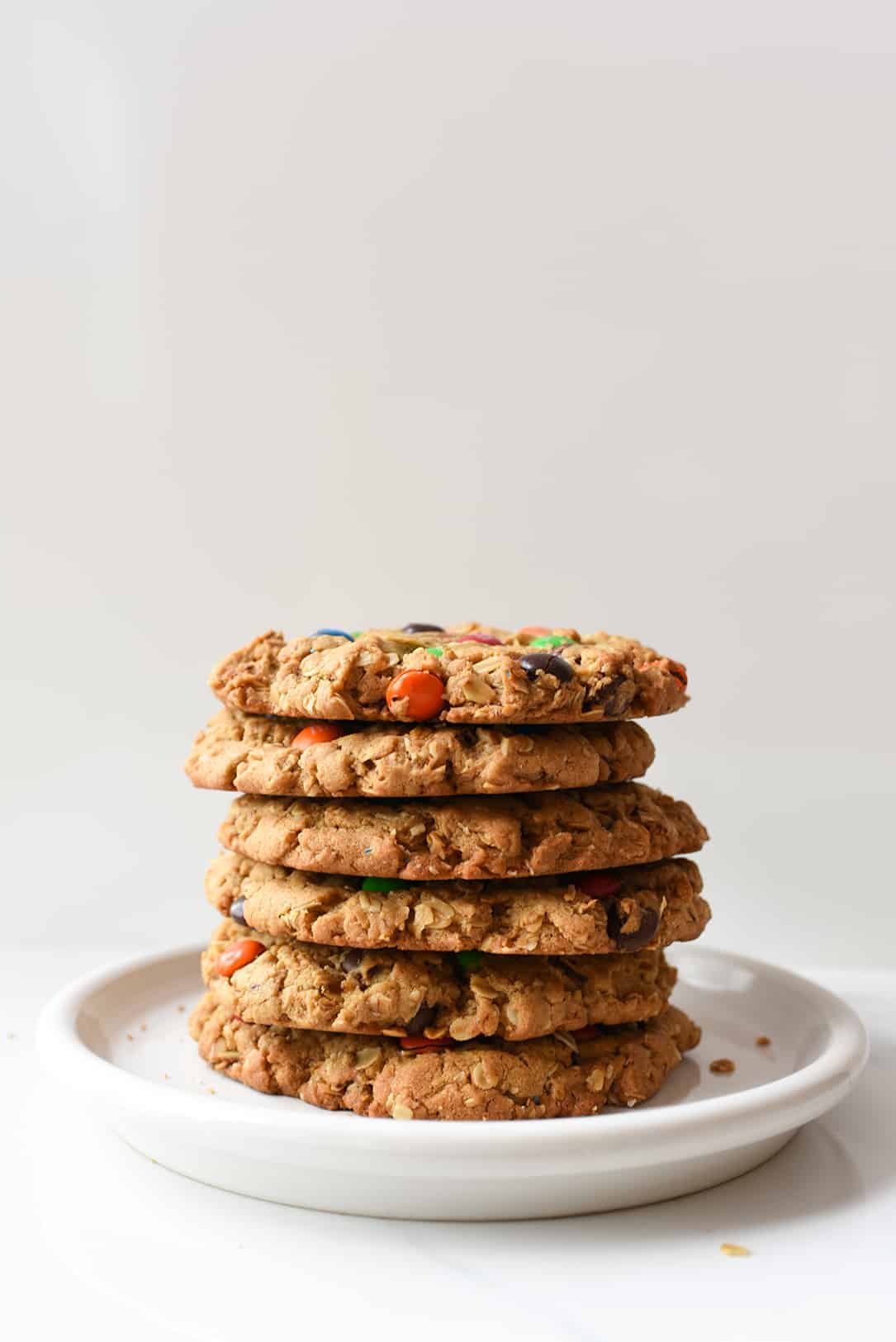 This classic recipe is kicked up a notch and made GIANT! Peanut butter and M&M's work together to create the best cookie recipe ever! #monstercookies #cookies #cookierecipe #iambaker Giant Monster Cookies stacked on White Plate