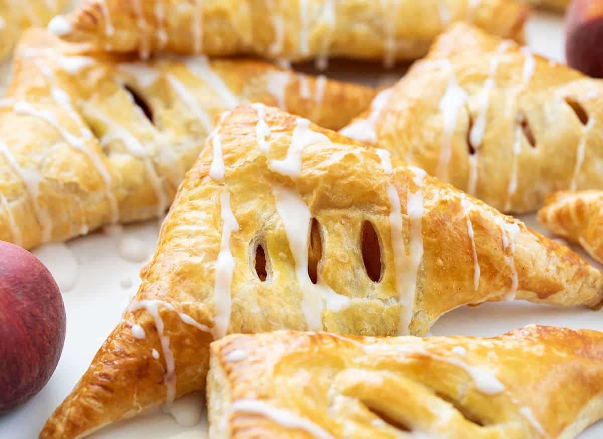 Peach Turnovers are squares of puff pastry filled with spiced canned peaches, baked to a golden brown, and drizzled with a sweet glaze. The flaky texture and fruity goodness will surely leave you craving more, and they are so easy to make! Enjoy a turnover as a breakfast treat, with a cup of coffee, or as a dessert. And, you can even make them in an air fryer! Peach Turnovers on a White Counter with Peaches.