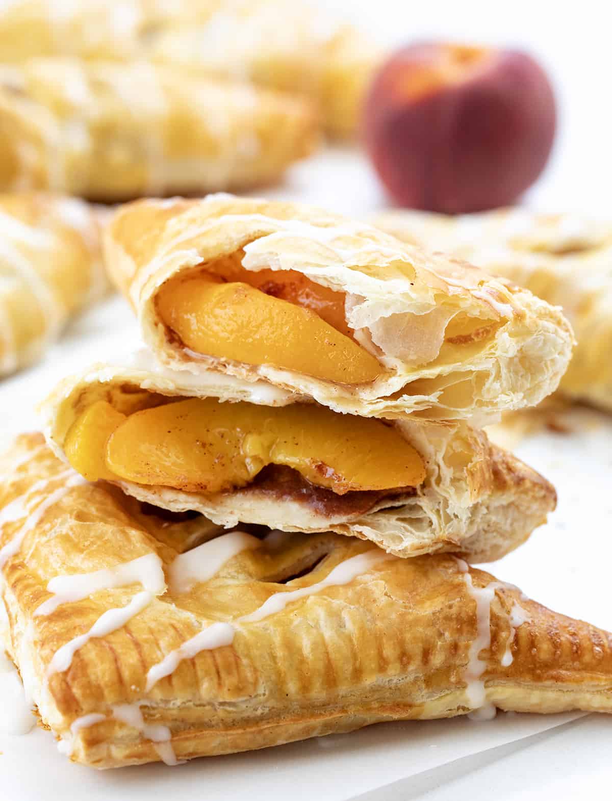 Peach Turnovers are squares of puff pastry filled with spiced canned peaches, baked to a golden brown, and drizzled with a sweet glaze. The flaky texture and fruity goodness will surely leave you craving more, and they are so easy to make! Enjoy a turnover as a breakfast treat, with a cup of coffee, or as a dessert. And, you can even make them in an air fryer! A Peach Turnovers Torn in Half Showing Peaches Inside.