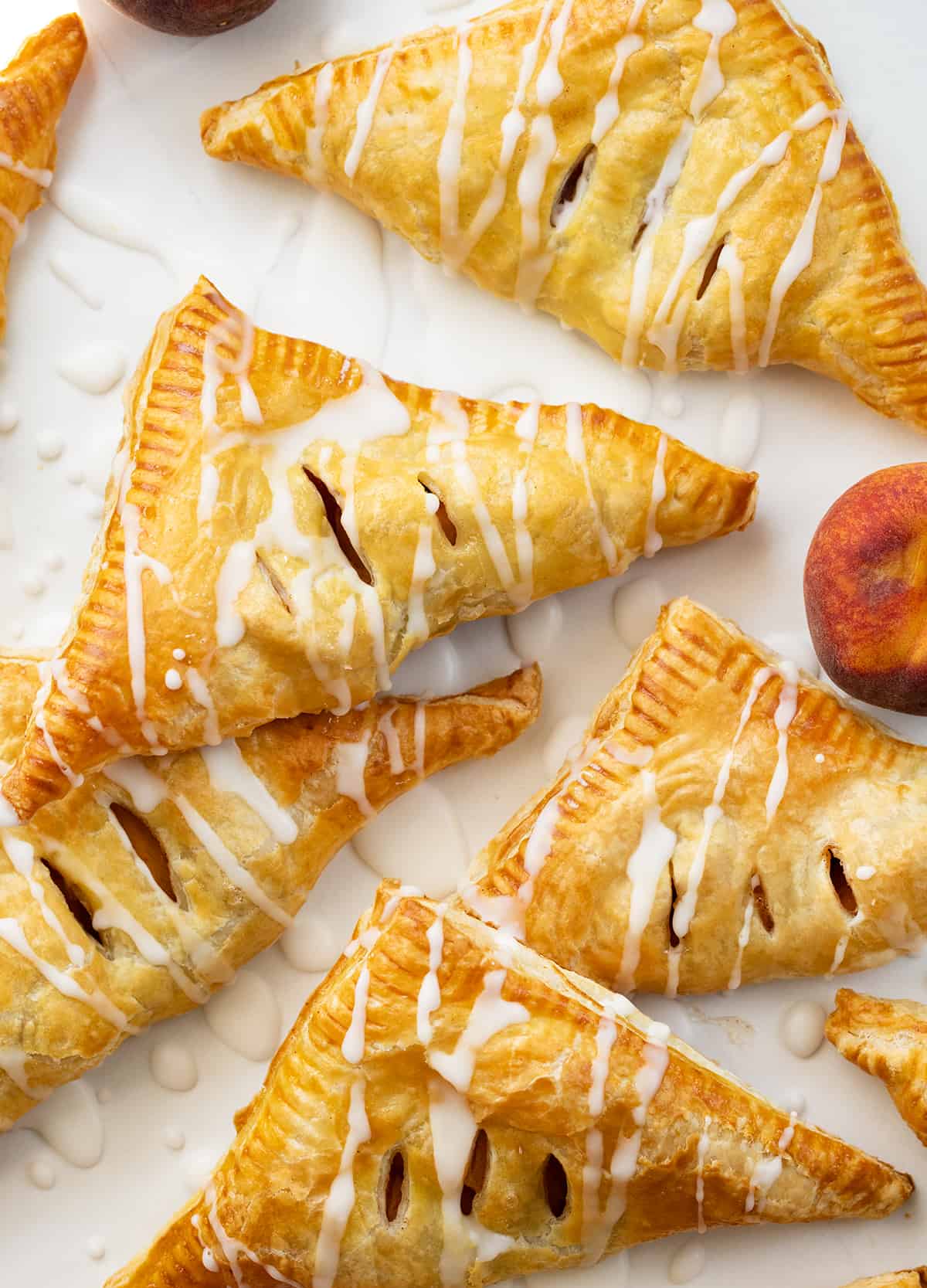 Peach Turnovers are squares of puff pastry filled with spiced canned peaches, baked to a golden brown, and drizzled with a sweet glaze. The flaky texture and fruity goodness will surely leave you craving more, and they are so easy to make! Enjoy a turnover as a breakfast treat, with a cup of coffee, or as a dessert. And, you can even make them in an air fryer! Peach Turnovers on a Piece of White Parchment with Peaches.