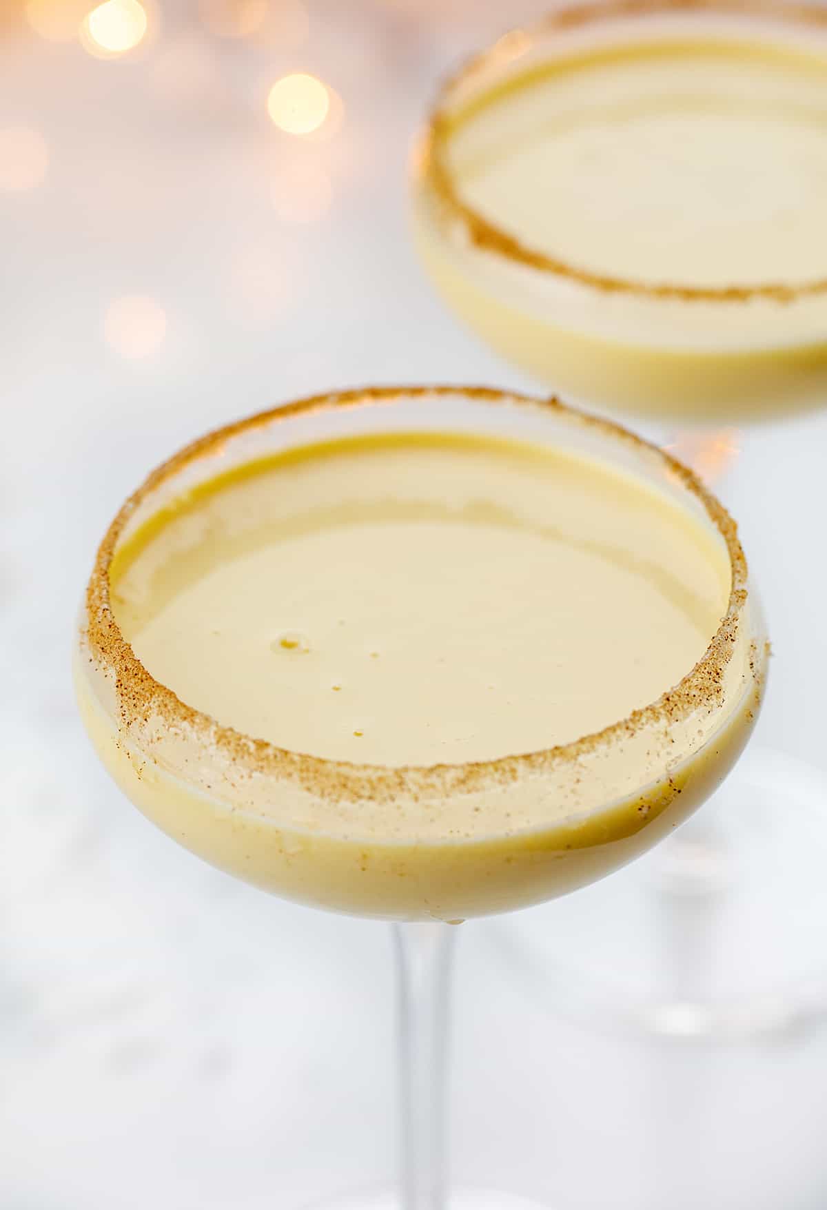 Rumchata Eggnog is an alcoholic beverage made with eggnog and RumChata® served in a glass with a cinnamon-sugar rim. It's the perfect cocktail to serve at your Christmas and New Year's Eve parties! Glasses of RumChata Eggnog in Front of Twinkling Lights.