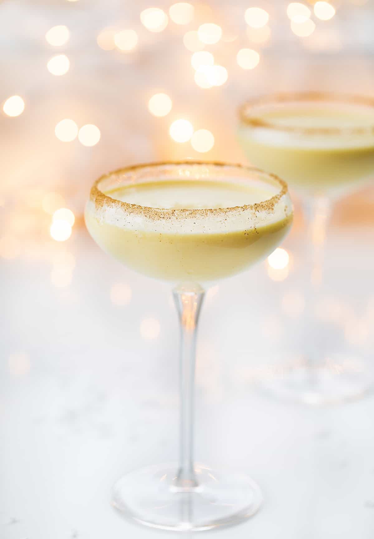 Rumchata Eggnog is an alcoholic beverage made with eggnog and RumChata® served in a glass with a cinnamon-sugar rim. It's the perfect cocktail to serve at your Christmas and New Year's Eve parties! Glasses of RumChata Eggnog in Front of Twinkling Lights.